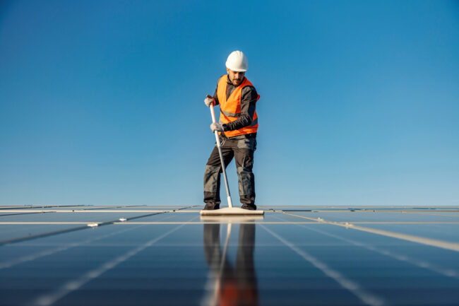 A handyman brushing solar panels with broom on the roof.